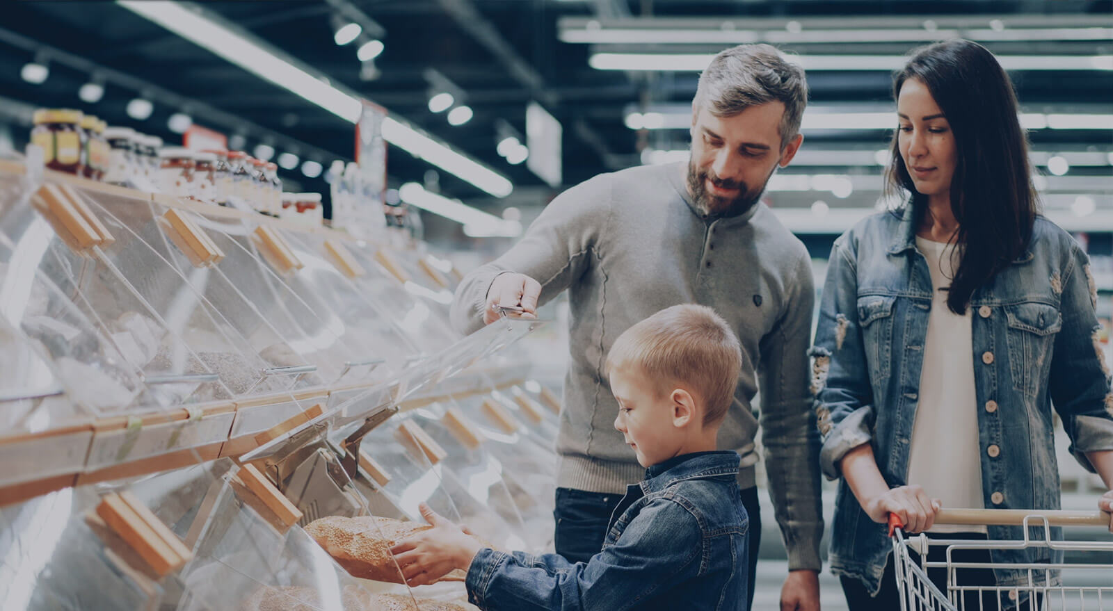 A family of four shopping together in a brightly lit supermarket aisle, examining products and filling their cart.