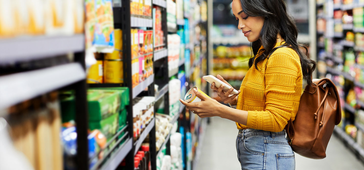 A woman examines a food item while shopping in a grocery store aisle.