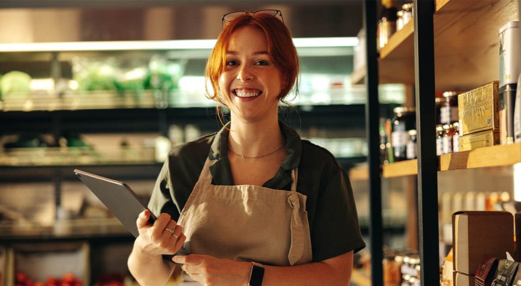 A woman in an apron holds a tablet, looking engaged in a grocery store.